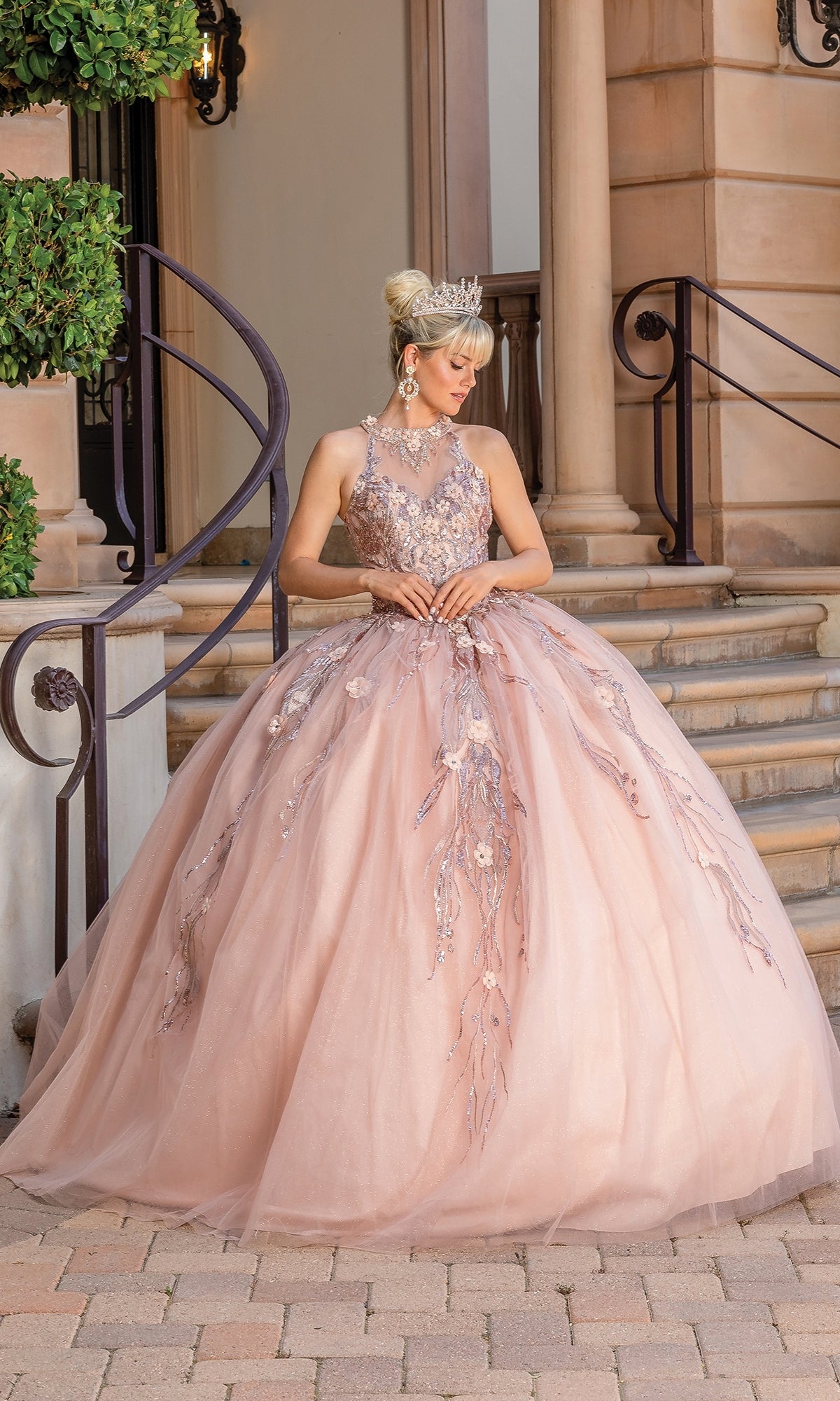 A woman in the Sleeveless Quinceanera Dress 1677, a pink ball gown with floral embroidery and a tiara, stands on steps before a columned building.